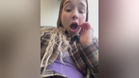 Media: A video of a young woman with fair skin, light brown hair, and blue eyes, wearing a plaid shirt, eating a lollipop with a shocked expression, blurred background.