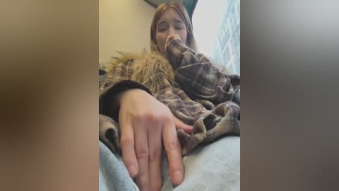 Media: Video of a young woman with light skin, straight brown hair, and a plaid shirt, lying on a bed, reaching out with her left hand. The background shows a blurred window with a curtain.