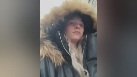 Media: A video shows a young Caucasian woman with light skin, wearing a large, dark fur-trimmed hooded coat and black earmuffs, gazing upwards. The background is blurred, with a muted color palette.