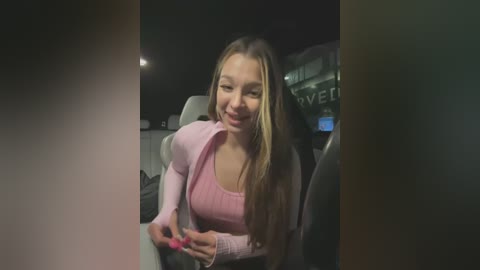 Media: Video of a smiling young woman with long blonde hair, wearing a pink sweater and pink nails, sitting in a dimly lit car with a \"VIP\" sign visible.