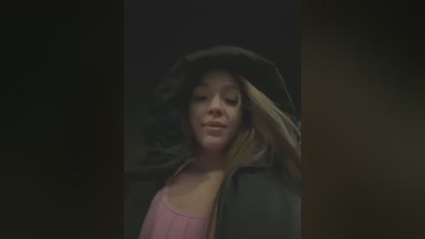 Media: Video of a young woman with fair skin and blonde hair, wearing a black hoodie and pink tank top, with a dark, blurred background.