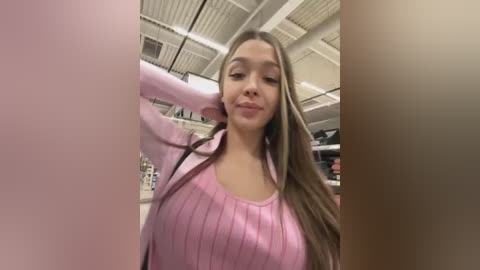 Media: Video of a young woman with long, straight brown hair, wearing a pink ribbed sweater, standing in a brightly lit, industrial-style room with visible ceiling panels and shelving in the background.