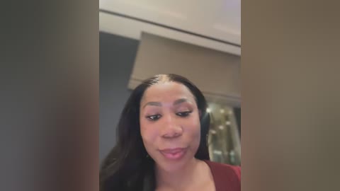 Media: Video of a young Black woman with medium-brown skin and long, straight black hair, wearing a maroon top, looking slightly to the right, in an indoor setting with neutral-toned walls and a glass door.