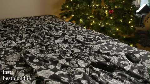 Media: A video of a bed covered with a black duvet featuring a white floral pattern, beside a decorated Christmas tree with warm, yellow lights and a red ornament, in a cozy indoor setting.