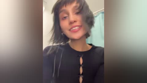 Media: Video of a young woman with short, dark hair and light brown skin, wearing a black top with a keyhole design, smiling in a bathroom with a blue shower curtain in the background.