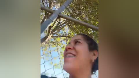 Media: Video of a woman with light brown hair, smiling upwards, looking through a chain-link fence at a tree canopy. The background shows dappled sunlight and green leaves.