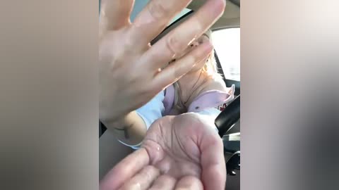 Media: Video of a blonde woman, in a light pink blouse, with a tattoo on her upper arm, driving a car. Her hands are on the steering wheel, and a man's hand is in the foreground, slightly blurred.