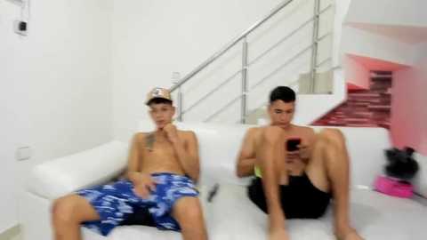 Media: Video of two shirtless, slim, young men in blue and black swim trunks, sitting on a white couch, looking at phones; modern, white minimalist living room background.