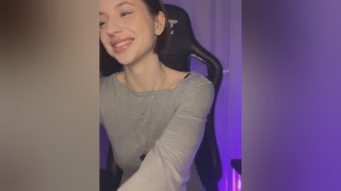 Media: Video of a smiling, light-skinned woman with short brown hair, wearing a gray long-sleeve top, sitting in a black gaming chair. Background includes purple lighting and a white curtain.