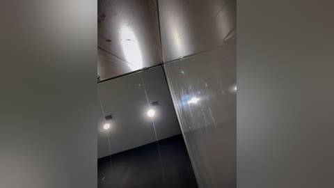 Media: Video of a modern, minimalist interior featuring a reflective ceiling with four recessed lights casting soft, diffuse light. The walls are painted a neutral, dark gray, and the ceiling is sleek and metallic, reflecting the lights and creating a visually dynamic, clean aesthetic.