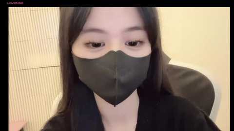 Media: Video of an East Asian woman with long black hair, wearing a black face mask and black dress, seated in a tiled bathroom.