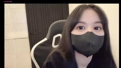 Media: Video of an Asian woman with long black hair, wearing a black face mask and black hoodie, sitting in a gaming chair against a beige wall and black vertical blinds.