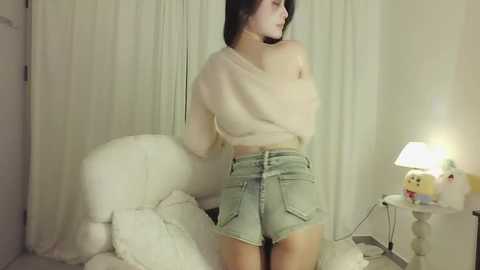 Media: Video of an Asian woman with long black hair, wearing a beige off-shoulder top and high-waisted denim shorts, standing in a softly lit room with white curtains, plush toys, and a table lamp.