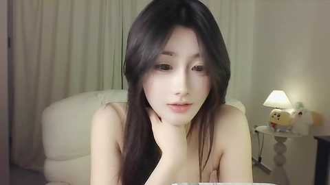 Media: Video of a young East Asian woman with long black hair, pale skin, and a delicate face, sitting in a softly lit bedroom with white curtains and a plush white chair.