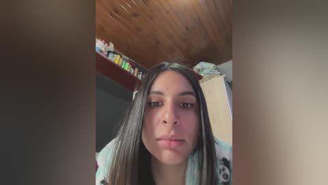 Media: Video of a young Latina woman with long dark hair, wearing a floral-patterned robe, standing in a wooden-paneled room with a shelf of colorful objects in the background.