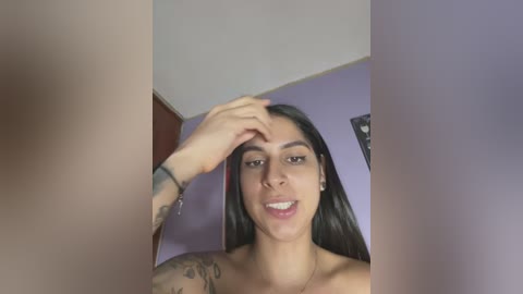 Media: Video of a Latina woman with long black hair, light skin, and tattoos on her arm, smiling, touching her forehead, in a bedroom with purple walls and framed art.