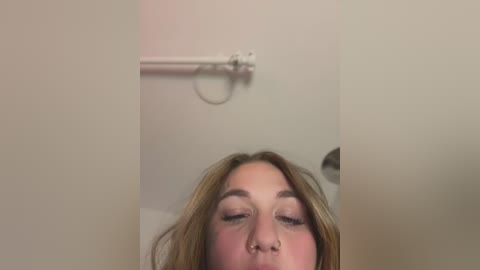 Media: A video of a young woman with light skin and long, light brown hair, eyes closed, leaning forward in a bathroom with a beige wall and a white towel rack.