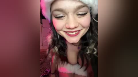 Media: Video of a young woman with fair skin and curly brown hair, wearing a Santa hat and red lipstick, smiling in a dimly lit room with a pinkish hue.