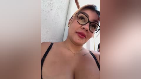 Media: A video of a Latina woman with short hair, medium skin tone, wearing black-framed glasses and a black bra. She has a tattoo on her shoulder and a necklace. The background shows a white wall and a window.