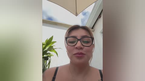 Media: A video of a plus-sized woman with glasses, wearing a black bra, standing under a beige umbrella on a patio. Her facial expression is neutral.