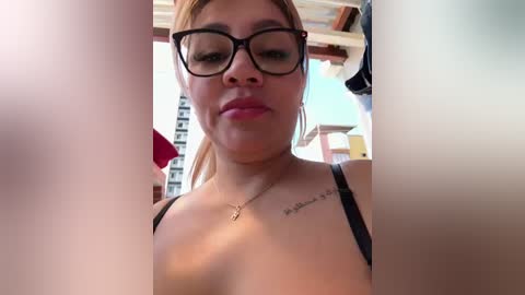 Media: Video of a young woman with light brown skin, wearing black-rimmed glasses, a black bra, and a necklace. She has a tattoo on her chest and is indoors, possibly in a bar.