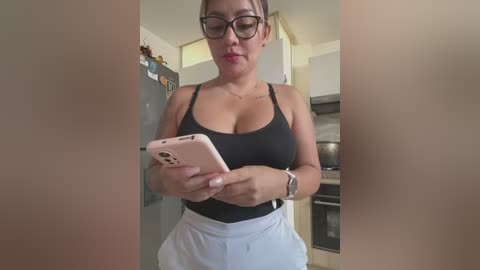 Media: Video of a curvy, light-skinned woman with glasses and a black tank top, standing in a modern kitchen, holding a pink smartphone.