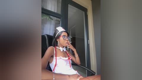 Media: A video of a slender, dark-skinned woman with long black hair, wearing a white nurse costume with red accents, sitting at a desk, smoking a cigarette.