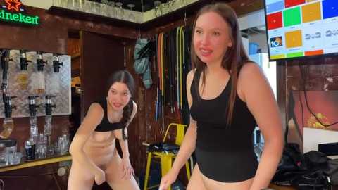 Media: Video of two young, slender, fair-skinned women with long brown hair, wearing black tank tops and no pants, in a cluttered, dimly lit bar with beer posters and glassware.