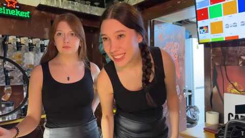 Media: Video of two young women with long brown hair in braids, wearing black tank tops and high-waisted leather skirts, standing in a cozy bar with beer taps, a beer sign, and colorful menu board.