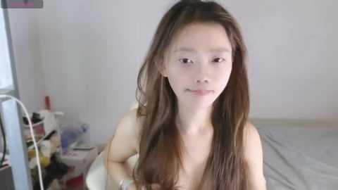 Media: Video of an Asian woman with long, straight brown hair, fair skin, and small breasts, sitting on a bed in a sparsely furnished room with a white wall, a medical device on the left, and a white blanket.