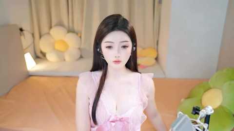 Media: A video of a young Asian woman with fair skin and long dark hair, wearing a pink lace lingerie top, standing in a softly lit room with beige curtains, white balloons, and a green cushion.