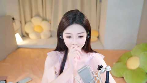 Media: A video of a young East Asian woman with long black hair, wearing a pink dress, listening to earphones, in a softly lit, cozy room with floral decor and a large green cushion.