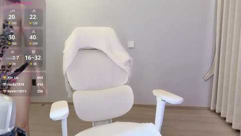 Media: A video of a modern, white ergonomic chair with a draped towel in a minimalist room with light walls and beige curtains.