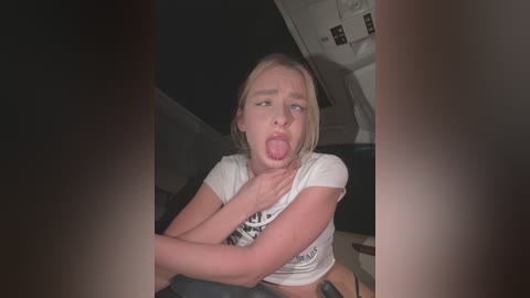 Media: A video captures a young blonde woman with fair skin, wearing a white T-shirt, sticking out her tongue and touching her neck, sitting in a dimly lit car interior.