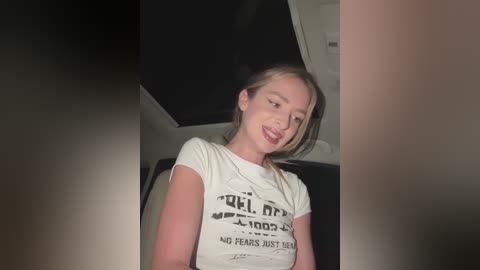 Media: Video of a young Caucasian woman with fair skin, blonde hair, wearing a white t-shirt with black text. She has a playful smile and is indoors, likely a bedroom, with dim lighting.