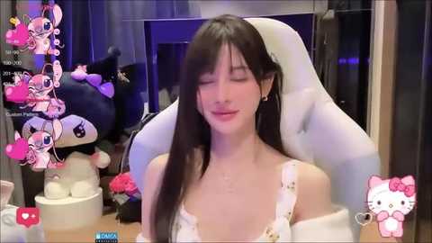 Media: Video of a smiling, light-skinned Asian woman with long black hair, wearing a white off-shoulder dress, sitting in a white gaming chair. Background features plush toys and a soft pink and purple color scheme.