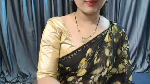 Media: Video of a South Asian woman with light skin, wearing a gold blouse under a black sari with yellow floral patterns. She has short, dark hair and wears a delicate necklace. Gray, silky fabric drapes in the background.