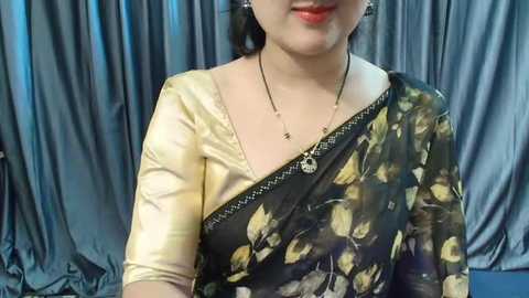 Media: Video of a South Asian woman in a black floral sari with a golden border, wearing a gold dupatta and silver earrings, against gray satin curtains.