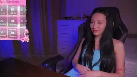 Media: A video of a fair-skinned woman with long black hair, wearing a blue dress, sitting in a black gaming chair. She looks at a pink and blue screen displaying a leaderboard.