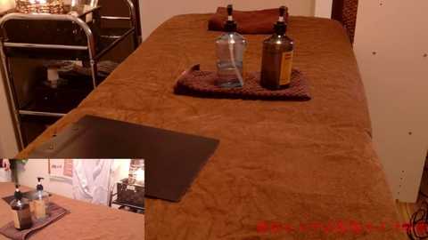 Media: Video of a rustic, wooden massage table covered with a brown, textured blanket. On the table are a glass bottle, a brown bottle, and a metal tray. The background includes a dark wooden chair and a partially visible kitchen area.