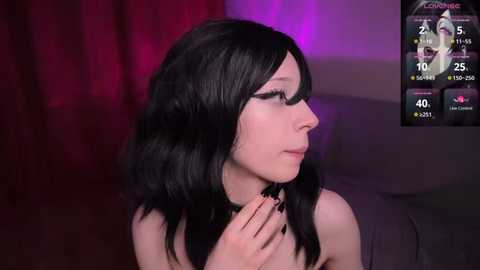 Video of a fair-skinned woman with long black hair, wearing a black choker, standing in a dimly lit room with red and purple lighting. She has a slender build and is looking to her left.