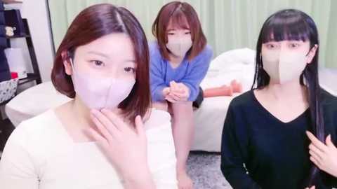 Media: Video of three East Asian women in a living room, wearing surgical masks, with one sitting on a white couch. They have short hair and are dressed in casual clothing.