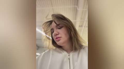 Media: Video of a young woman with shoulder-length brown hair, wearing a white robe, standing in a dimly lit room with exposed ceiling beams. She looks down, her expression neutral.