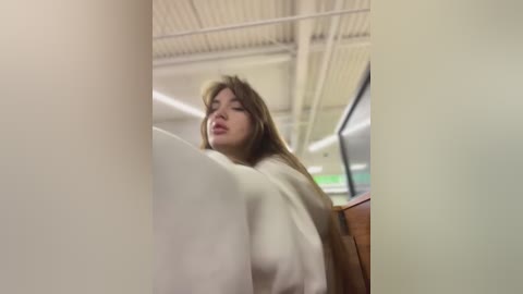 Media: Video of a young woman with long, light brown hair, wearing a white shirt, leaning against a white wall in a modern, well-lit indoor setting.