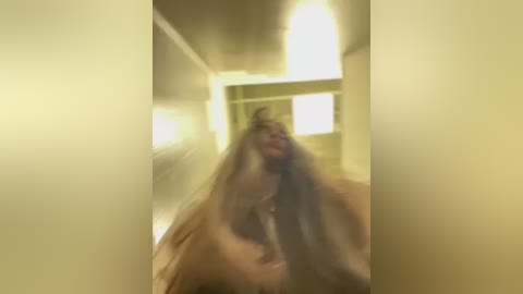 Media: A blurry video of a person with long, disheveled hair running down a corridor, bathed in yellowish light from overhead fixtures. The background shows a beige wall and a partially visible doorway.