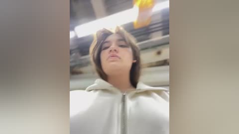 Media: Video of a woman with short, dark hair, wearing a white hoodie, looking down in an industrial setting with metal beams and fluorescent lights overhead.