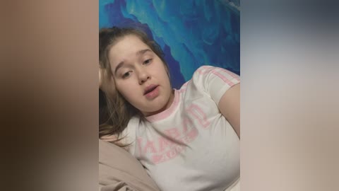 Media: Video of a young Caucasian woman with light brown hair, wearing a white t-shirt with pink text, lying on a bed with blue floral-patterned sheets, looking tired.