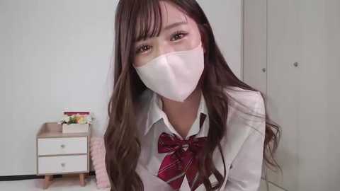 Media: Video of an Asian woman with long, wavy brown hair, wearing a white school uniform, red plaid tie, and white face mask, seated in a simple, white-walled room with a small wooden table and drawers.