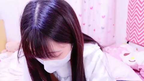 Media: Video of an Asian woman with long black hair and fair skin wearing a white mask, lying on a bed with pink and white patterned sheets, a stuffed animal, and a pink curtain with a heart pattern in the background.