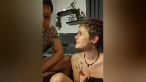 Media: Video of a tattooed man and a tattooed woman with short hair, both shirtless, sitting on a couch in a dimly lit room with a festive wreath on the wall.